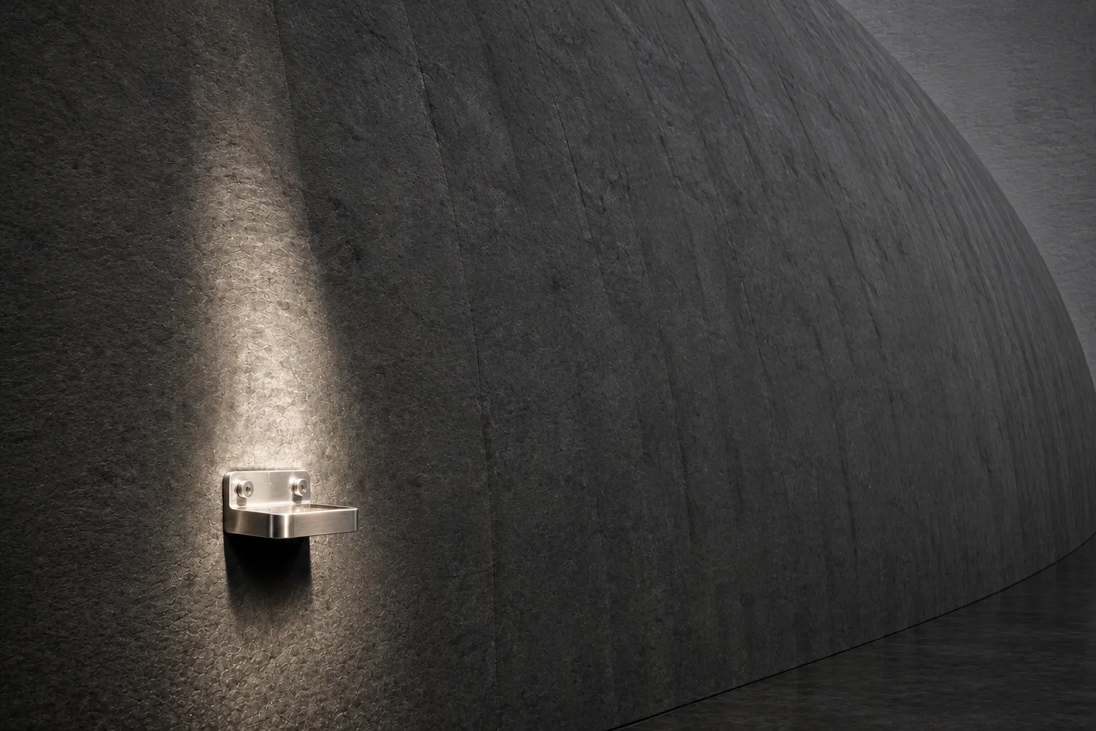 A small polished steel bracket bolted to a vast curved stone wall, lit by a single narrow beam while the wall stretches away into shadow.