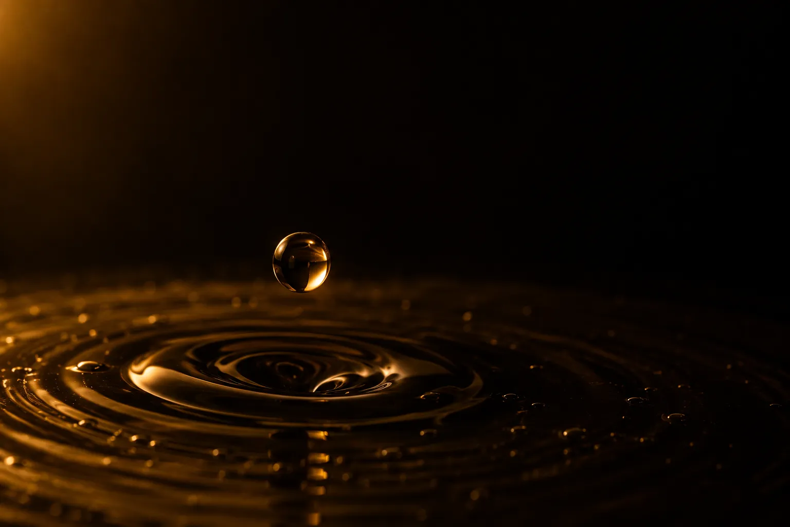 A single amber droplet falling towards a dark liquid surface caught mid-ripple, lit by one warm light against near darkness